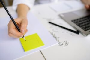 Close-up of hand sketching on a sticky note beside a laptop with paper clips on a white table.